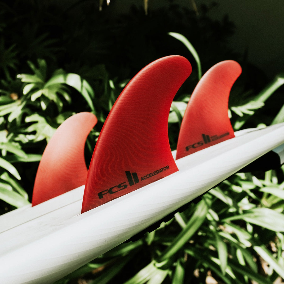 FCS II Accelerator Tri Fins - Eco Neo Glass: red fins on a board surrounded by tropical foliage