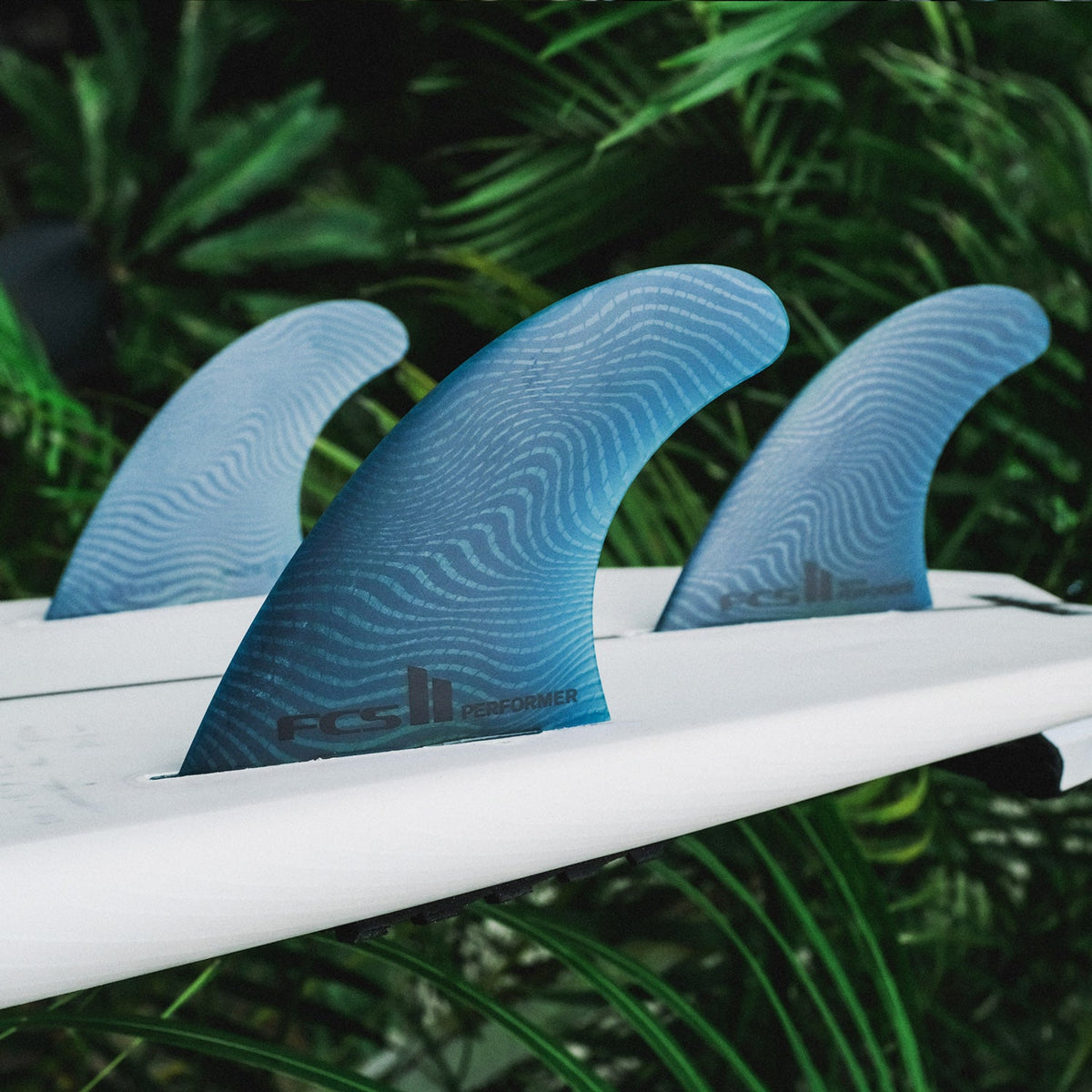 FCS II Performer Tri Fins - Eco Neo Glass: pacific blue thruster setup photographed beside tropical leaves