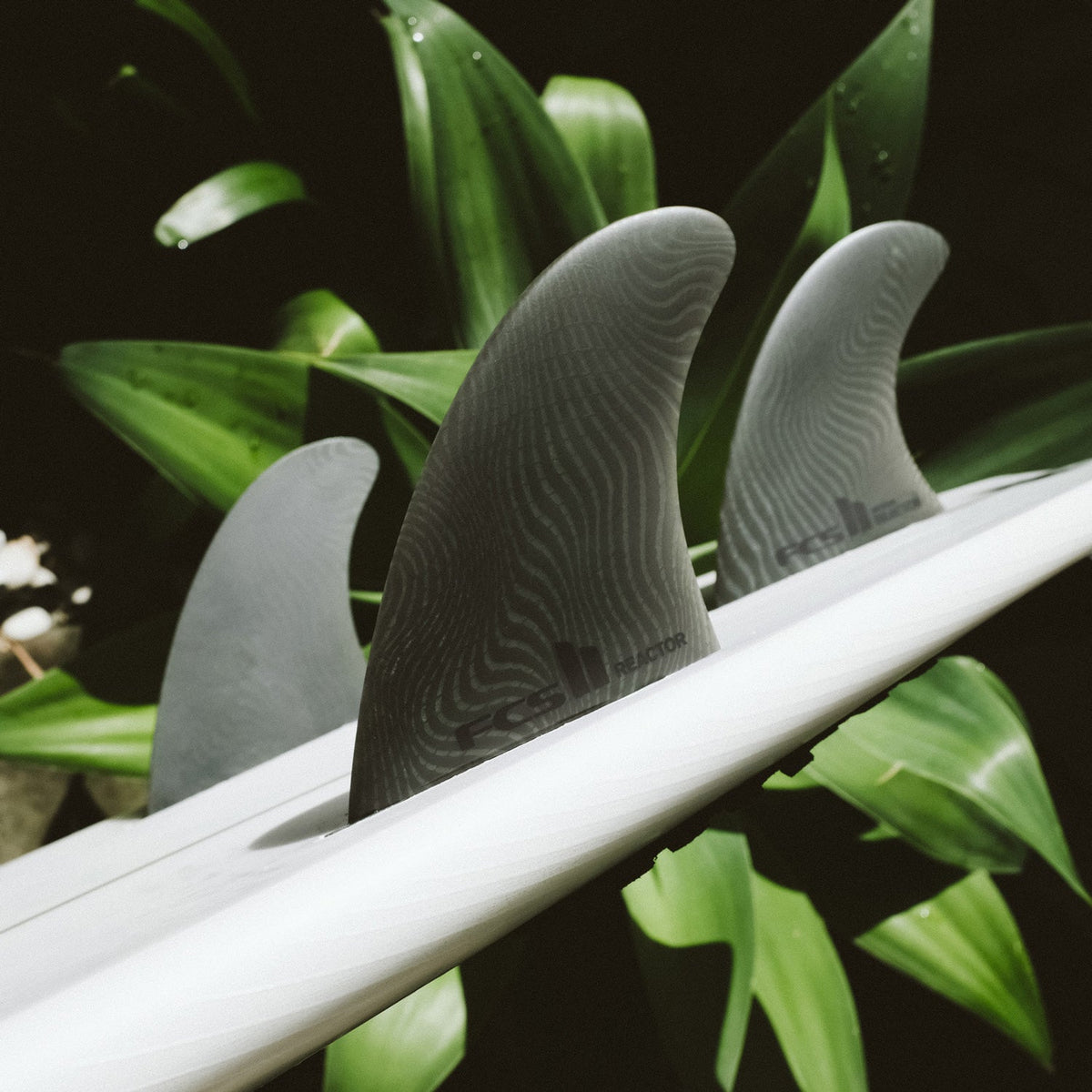 FCS II Reactor Tri Fins - Eco Neo Glass: reactor fins on a surfboard beside green foliage