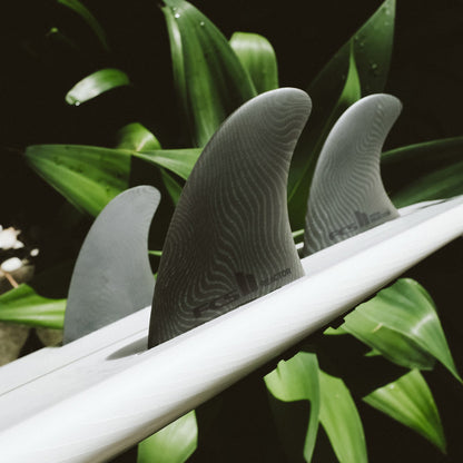 FCS II Reactor Tri Fins - Eco Neo Glass: reactor fins on a surfboard beside green foliage