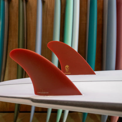 FCS II Christenson Twin - Performance Glass: blood Red twin fins mounted on a board among stored surfboards