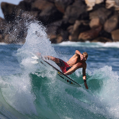 FCS II Mick Fanning Twin + Stabiliser Fins - PC + AirCore: surfer carving a wave near the rocky shoreline