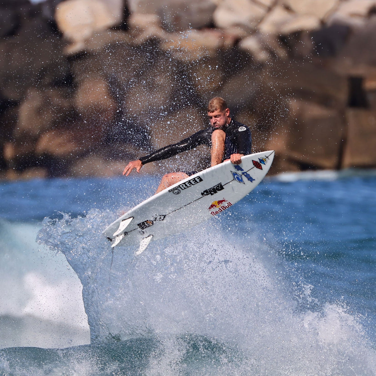 FCS II Mick Fanning Twin + Stabiliser Fins - PC + AirCore: surfer launching above a wave beside rocks