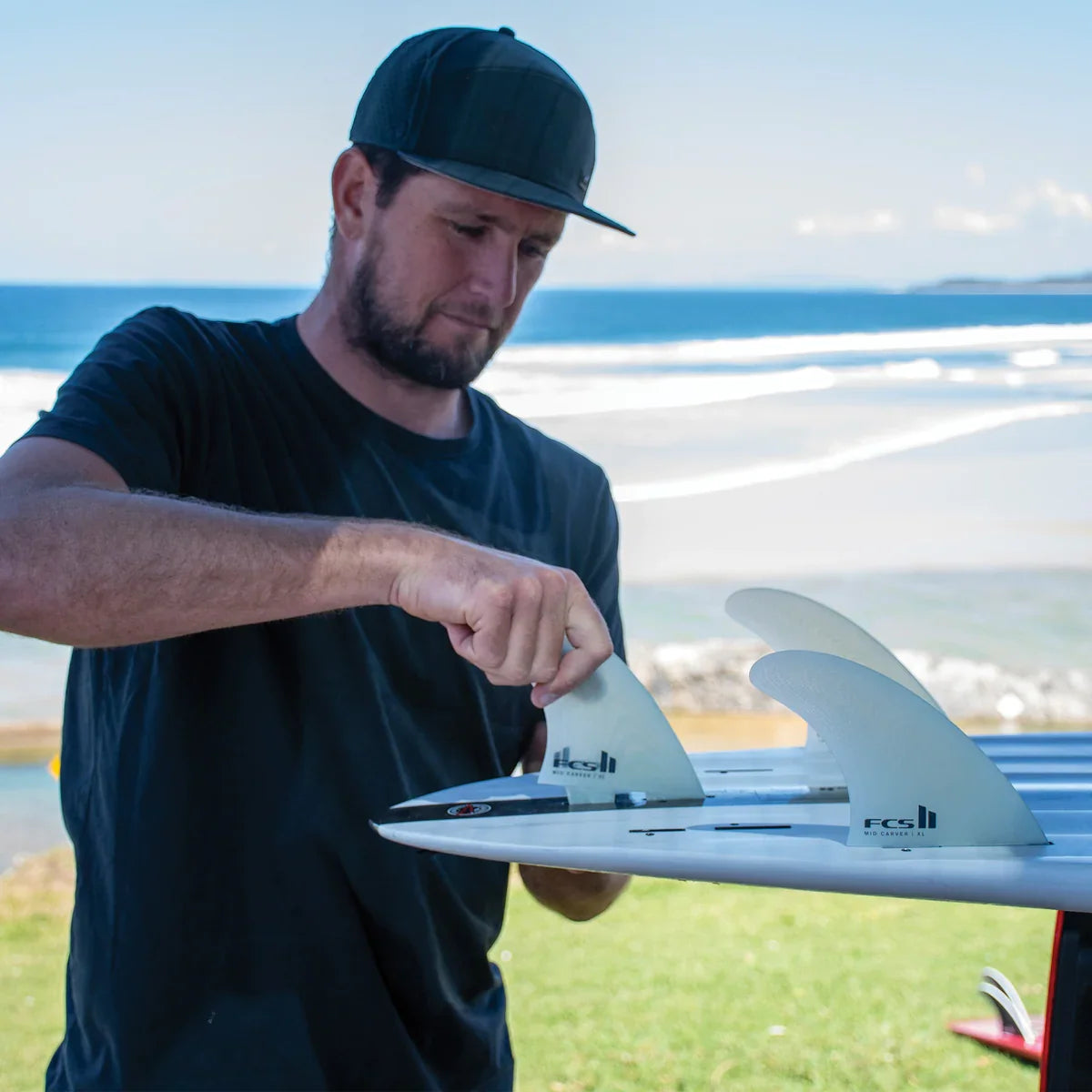FCS II Mid Carver 2 + 1 Fin Set - Performance Glass: man fitting a Clear fin to his board by the beach