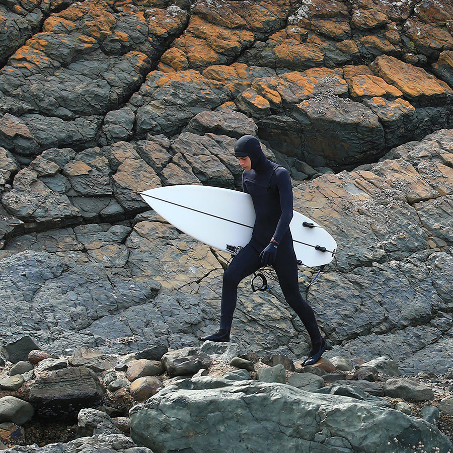 Ocean & Earth Free Flex Full Hood – 2 mm: Surfer carrying a board across rocks